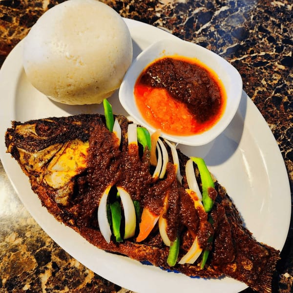 Whole fried fish with sauce, onions, and peppers, served with fufu and a side of red sauce.